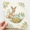 Baby kangaroo joey at a billabong with a wooden spoon, sweet potato and avocado on a gum-leaf plate.